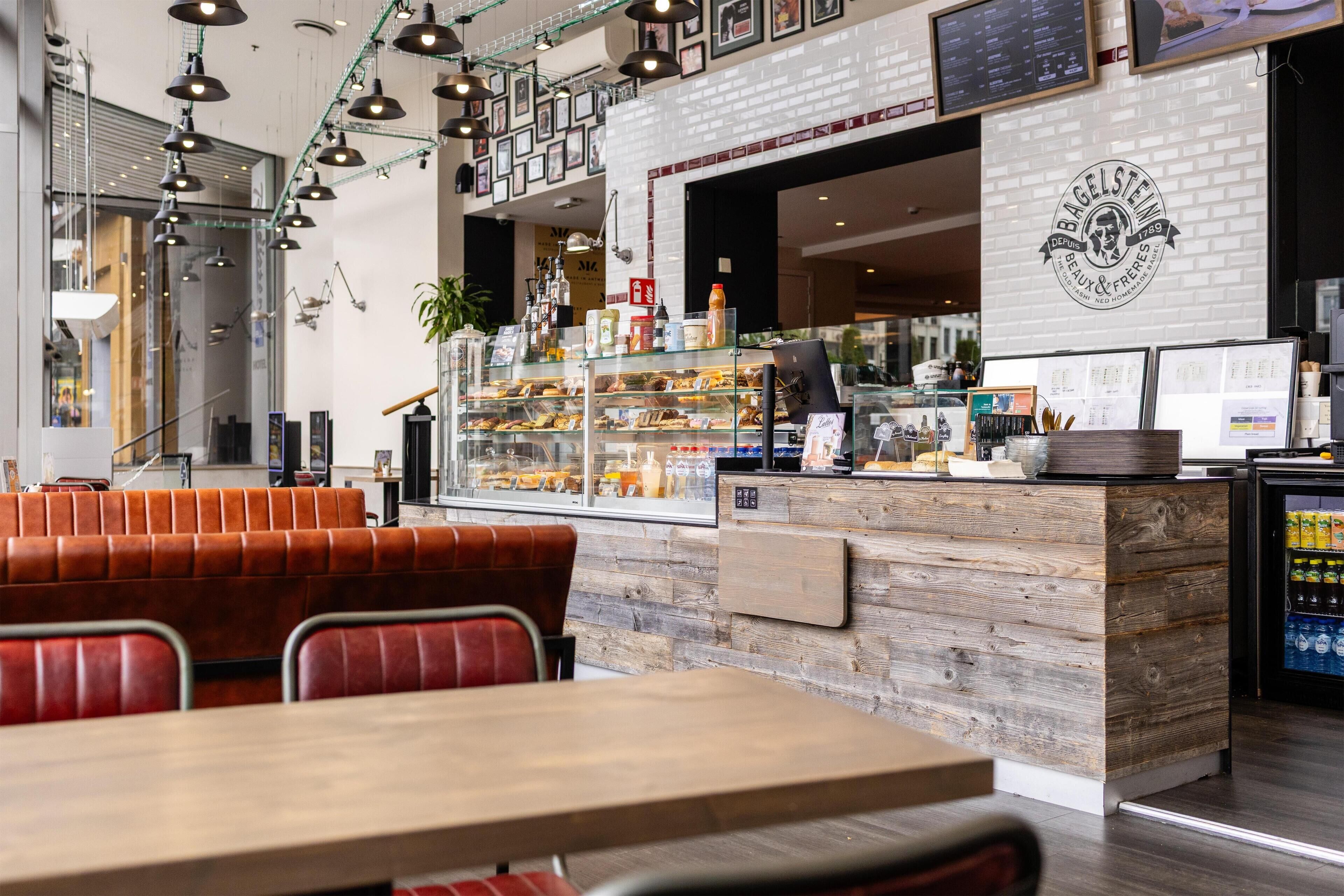 Interior de una cafetería con mesas de madera, asientos de cuero rojo, una vitrina con pasteles y una pizarra con el menú.