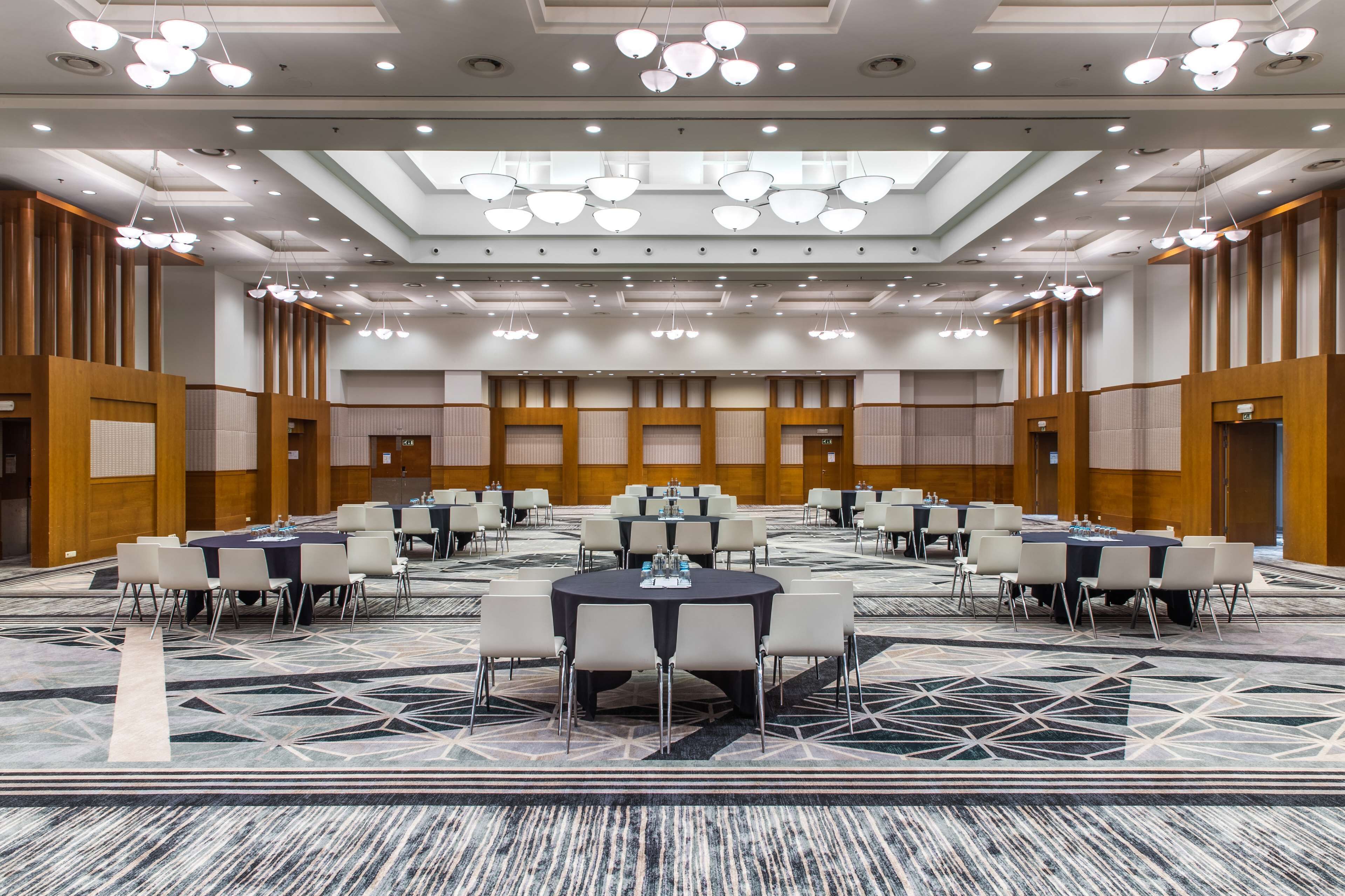 A large, well-lit conference room with multiple round tables and chairs, equipped with water dispensers.