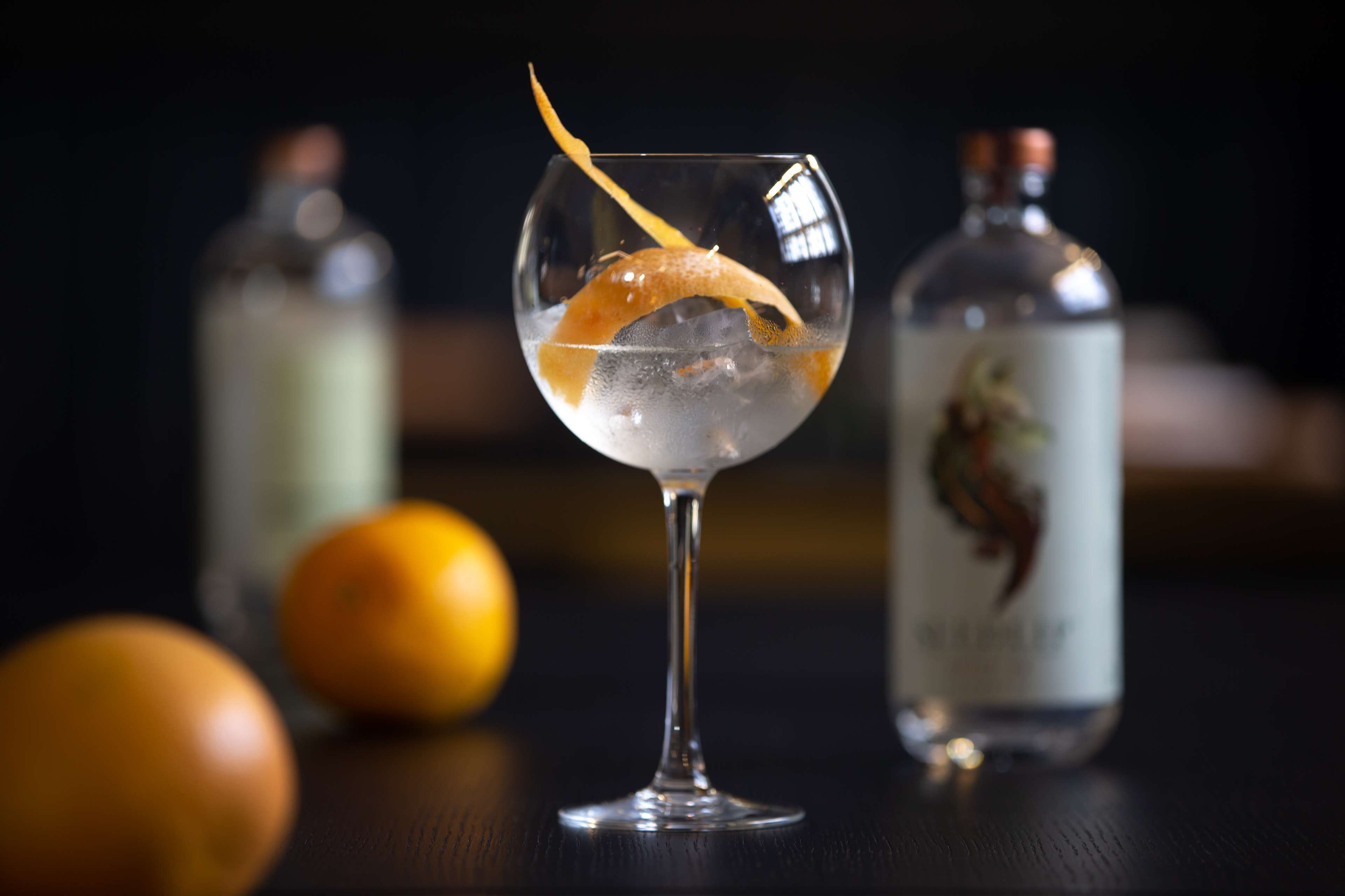A cocktail glass with an orange peel garnish, ice cubes, and two bottles in the background.