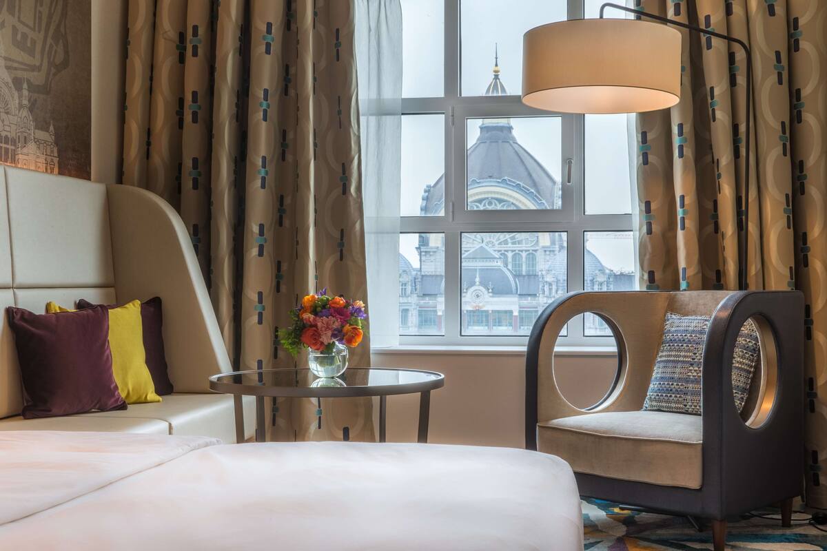 Una habitación de hotel con una cama, un sofá, una mesa redonda con un florero y una silla. Una ventana muestra vistas de un edificio con una cúpula.