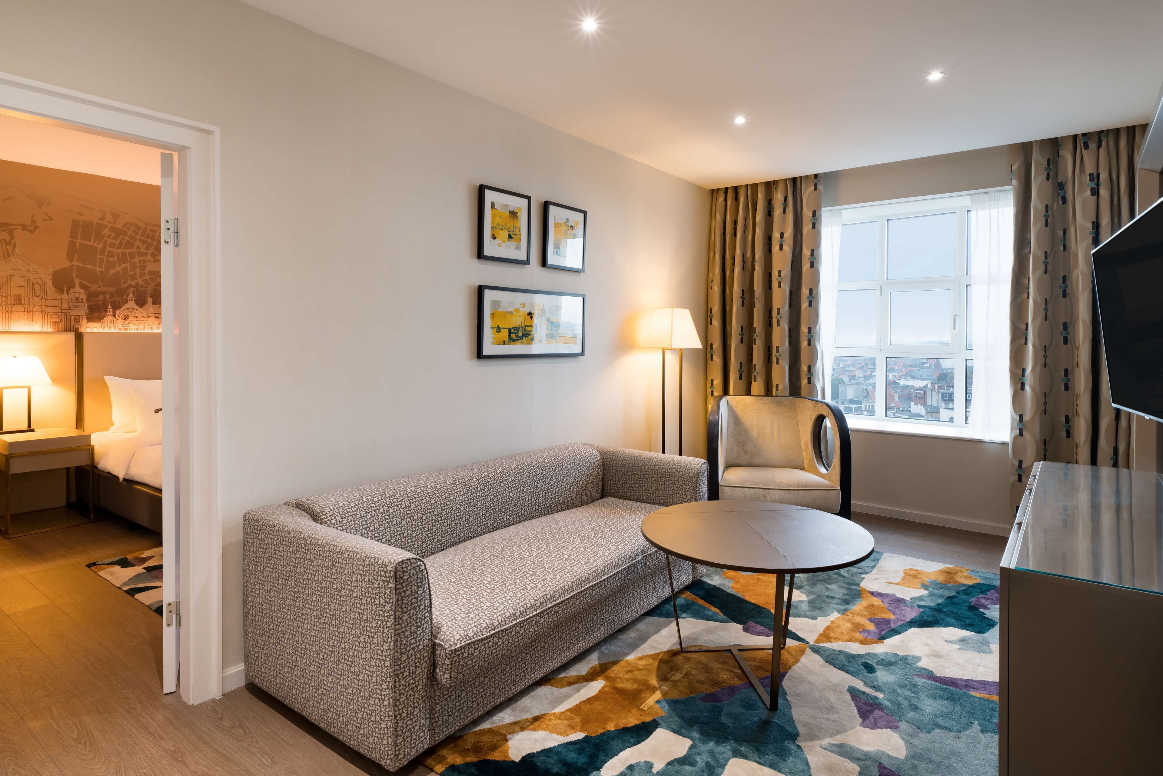 A hotel room with a sofa, a chair, a small table, a TV, and a window with curtains.
