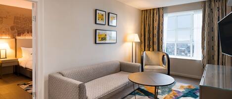 A hotel room with a sofa, a chair, a small table, a TV, and a window with curtains.