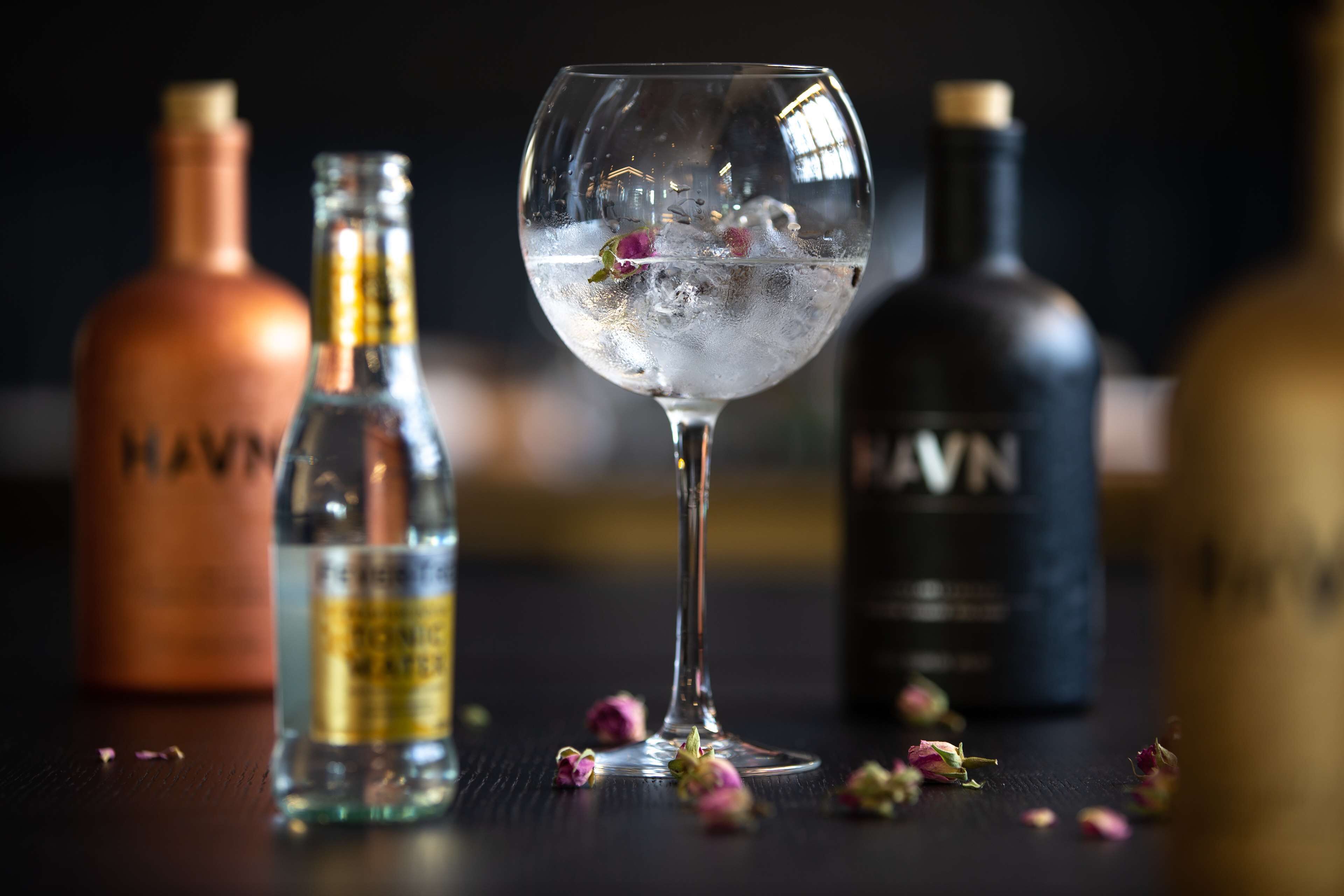 A glass of a clear liquid with ice cubes and a flower, surrounded by bottles of alcohol on a dark surface.