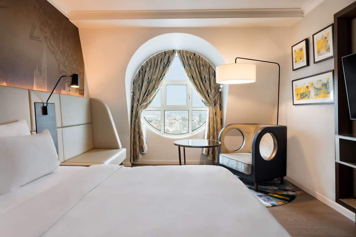Habitación de hotel con una cama grande, una mesa redonda, un sofá, un televisor y una ventana con cortinas.