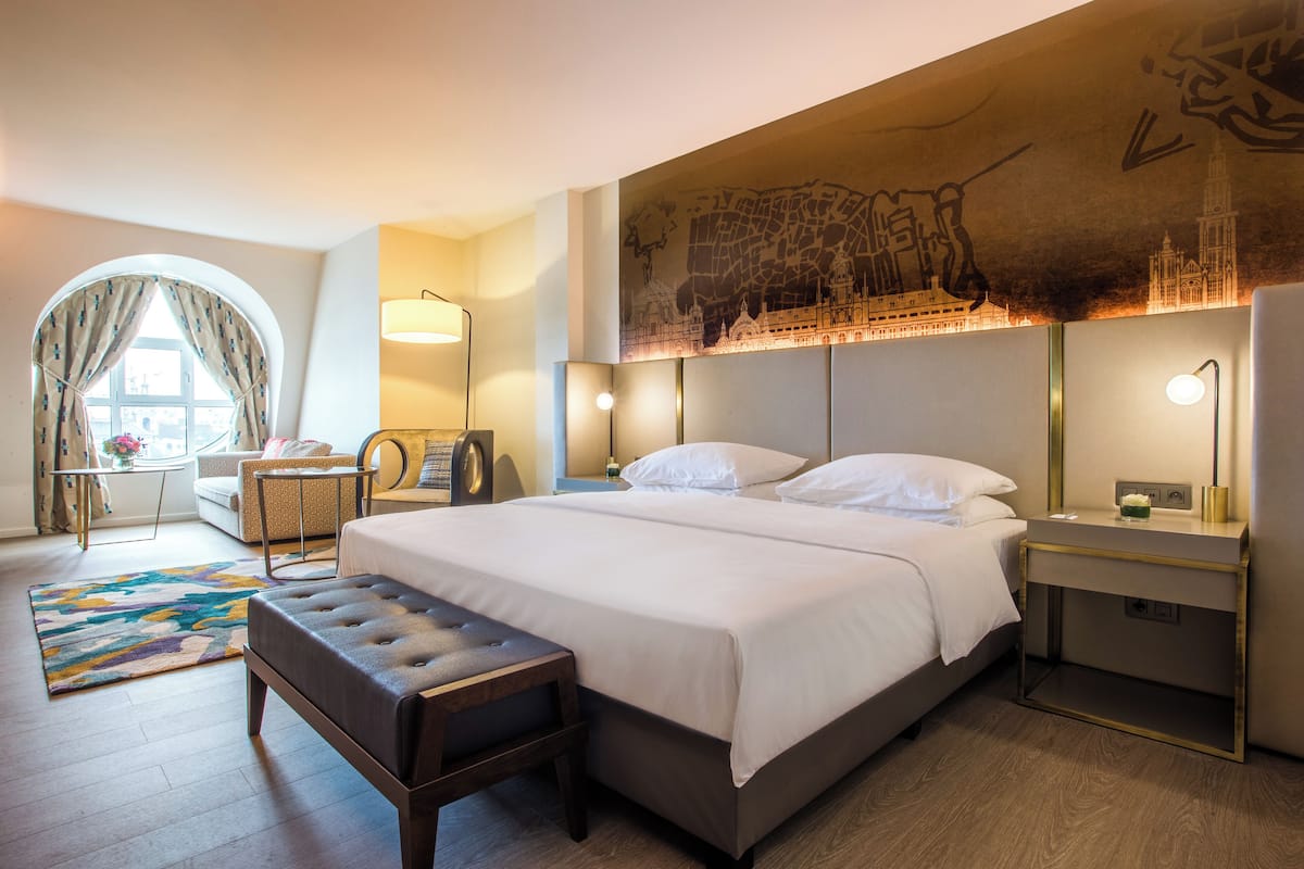 Habitación de hotel con una cama grande, una mesita de noche, una silla, un escritorio y un ventanal con cortinas.