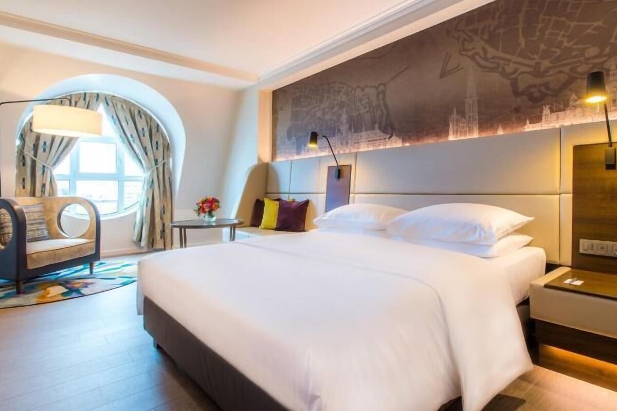 Una habitación de hotel moderna con una cama grande, una zona de estar con una silla, una mesita y una lámpara de pared.