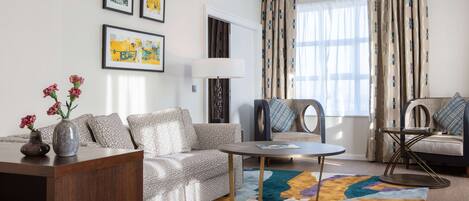 Uma sala de estar moderna com um sofá, poltronas, uma mesa de centro e um tapete. Quadros emoldurados adornam a parede e um abajur fica ao lado do sofá.