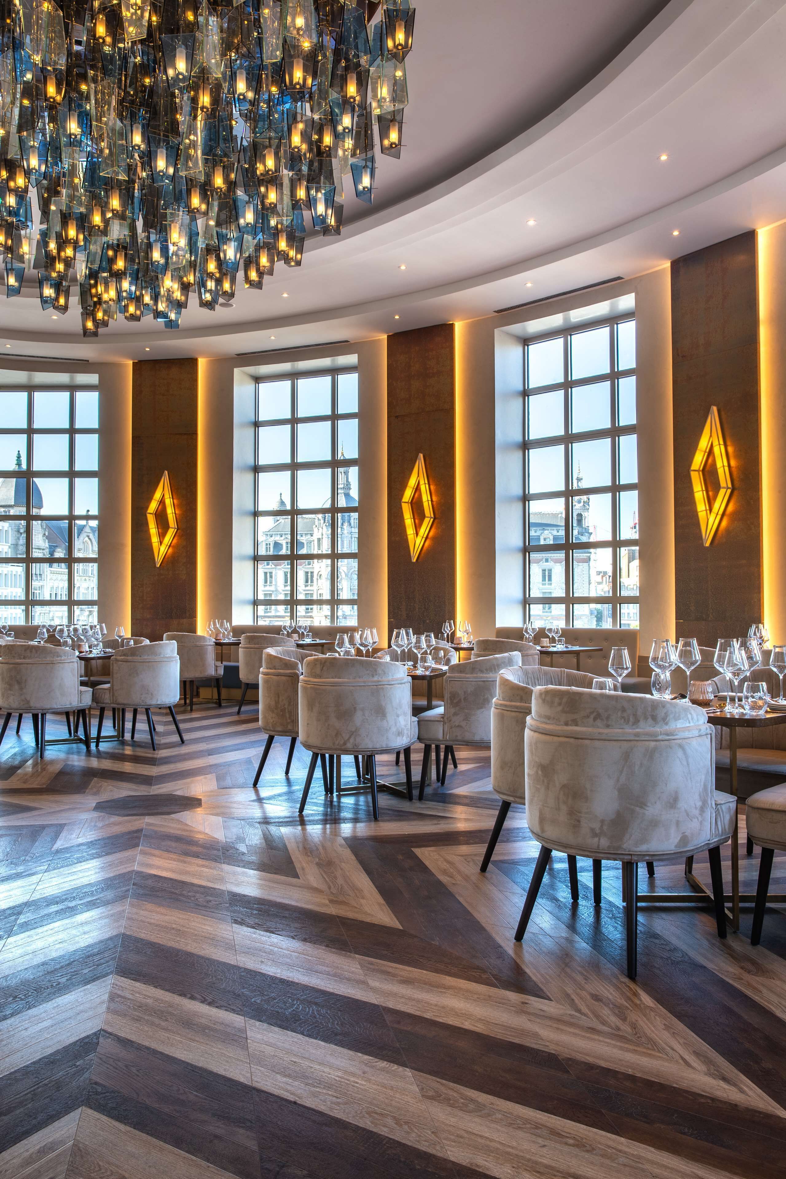 A modern dining area with a large chandelier, multiple tables with chairs, and large windows offering city views.