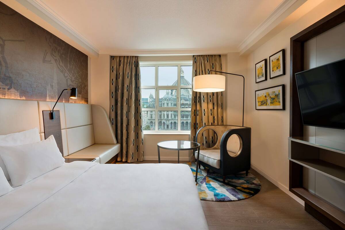 Habitación de hotel con cama, sofá, mesa redonda, silla, televisor y ventana con vistas a edificios.