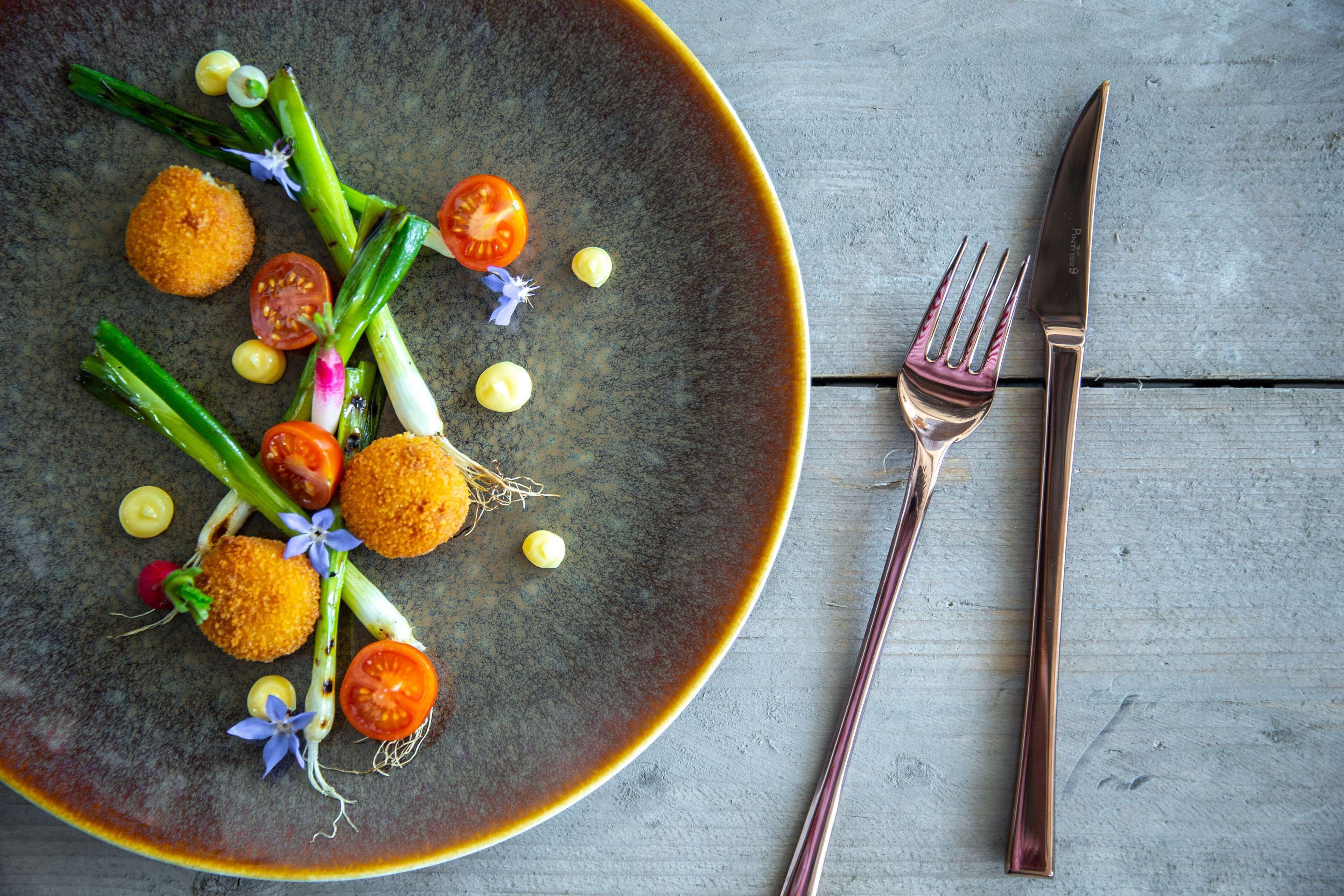 Une assiette de nourriture contenant des légumes et une croquette, accompagnée d’une fourchette et d’un couteau, posée sur une table en bois.