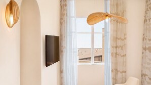 A modern living room with a flat-screen TV mounted on the wall, a round table, two white armchairs, and a large window with sheer curtains. - Hotel de l'Horloge (Avignon)