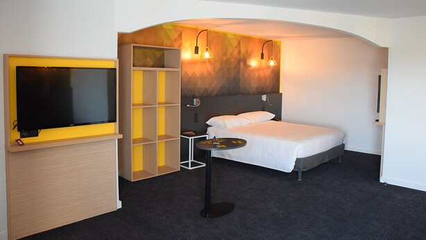 A hotel room with a bed, a TV, a small table, and a shelf unit. - ibis Styles Poitiers Nord (Poitiers)