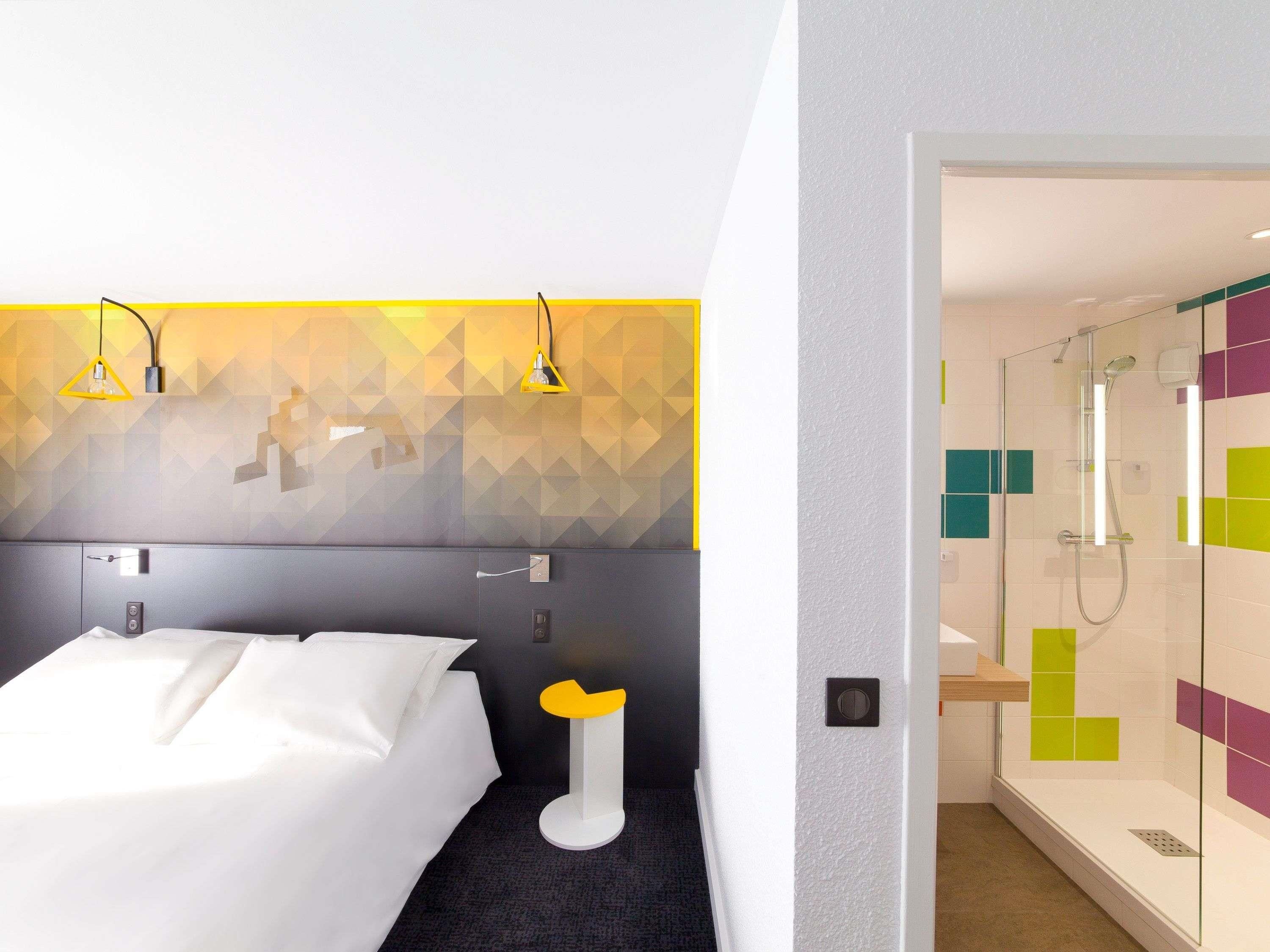 A hotel room with a bed, a bedside table, a shower area, and a wall with geometric patterns.