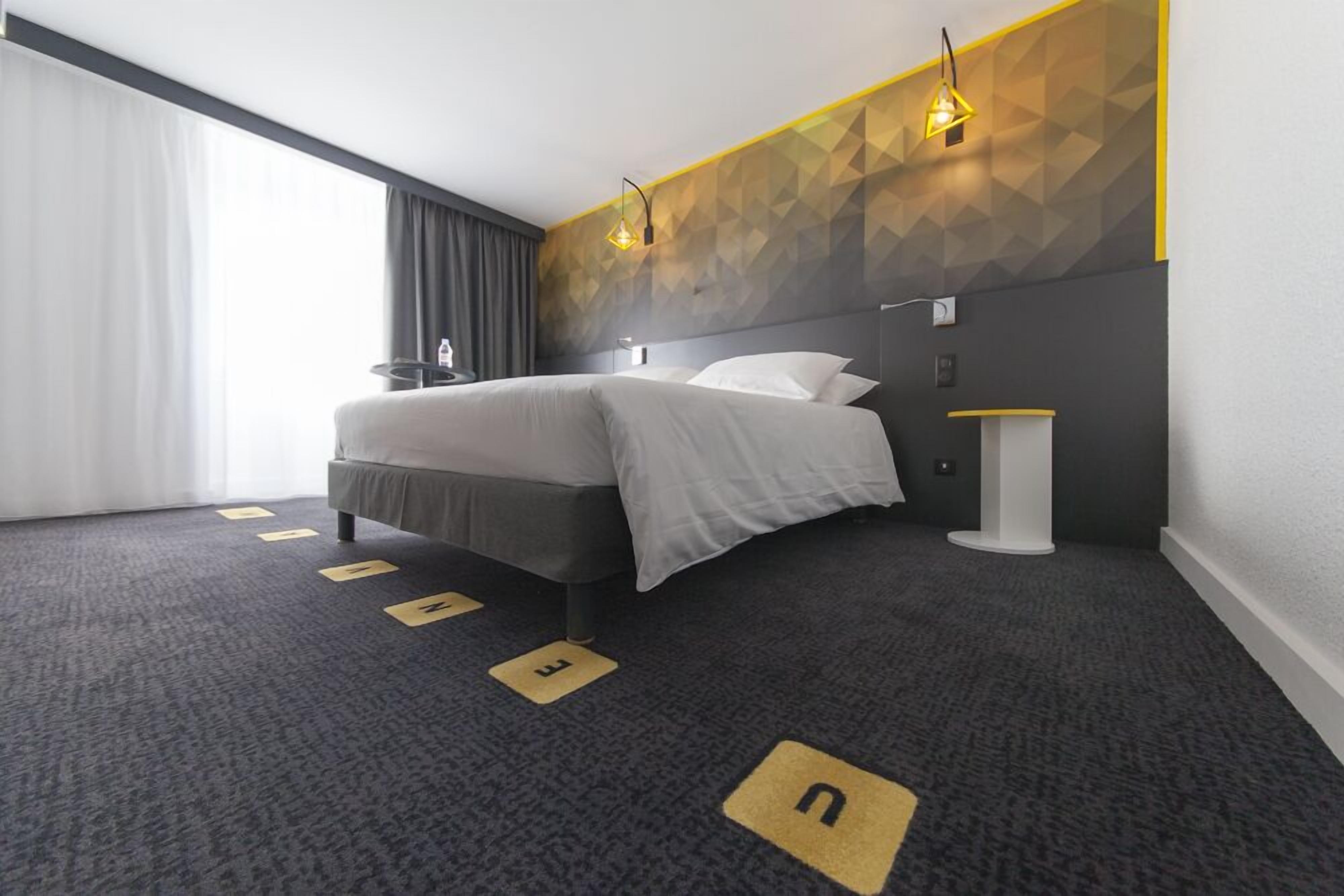 A modern hotel room with a large bed, a bedside table, a lamp, and a geometric-patterned wall.