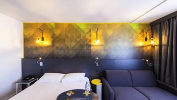 A modern hotel room with a bed, a sofa, a desk, and a wall with geometric patterns.