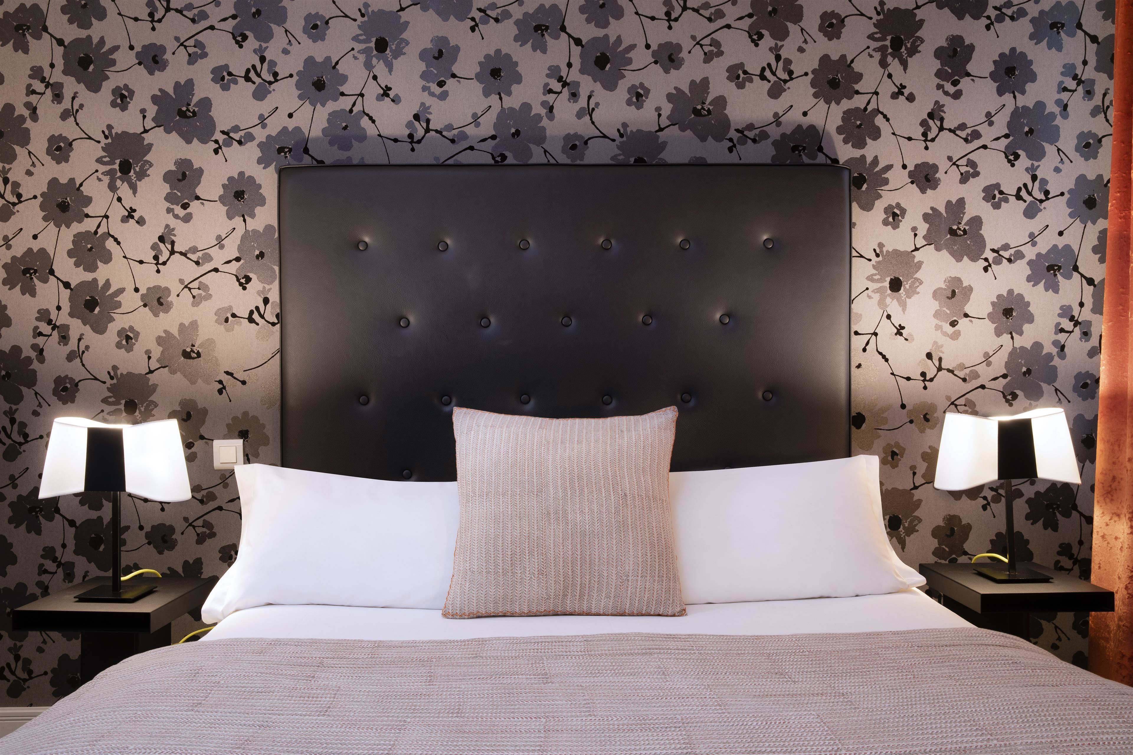 A neatly made bed with a dark headboard, two bedside lamps, and a floral wallpaper pattern.