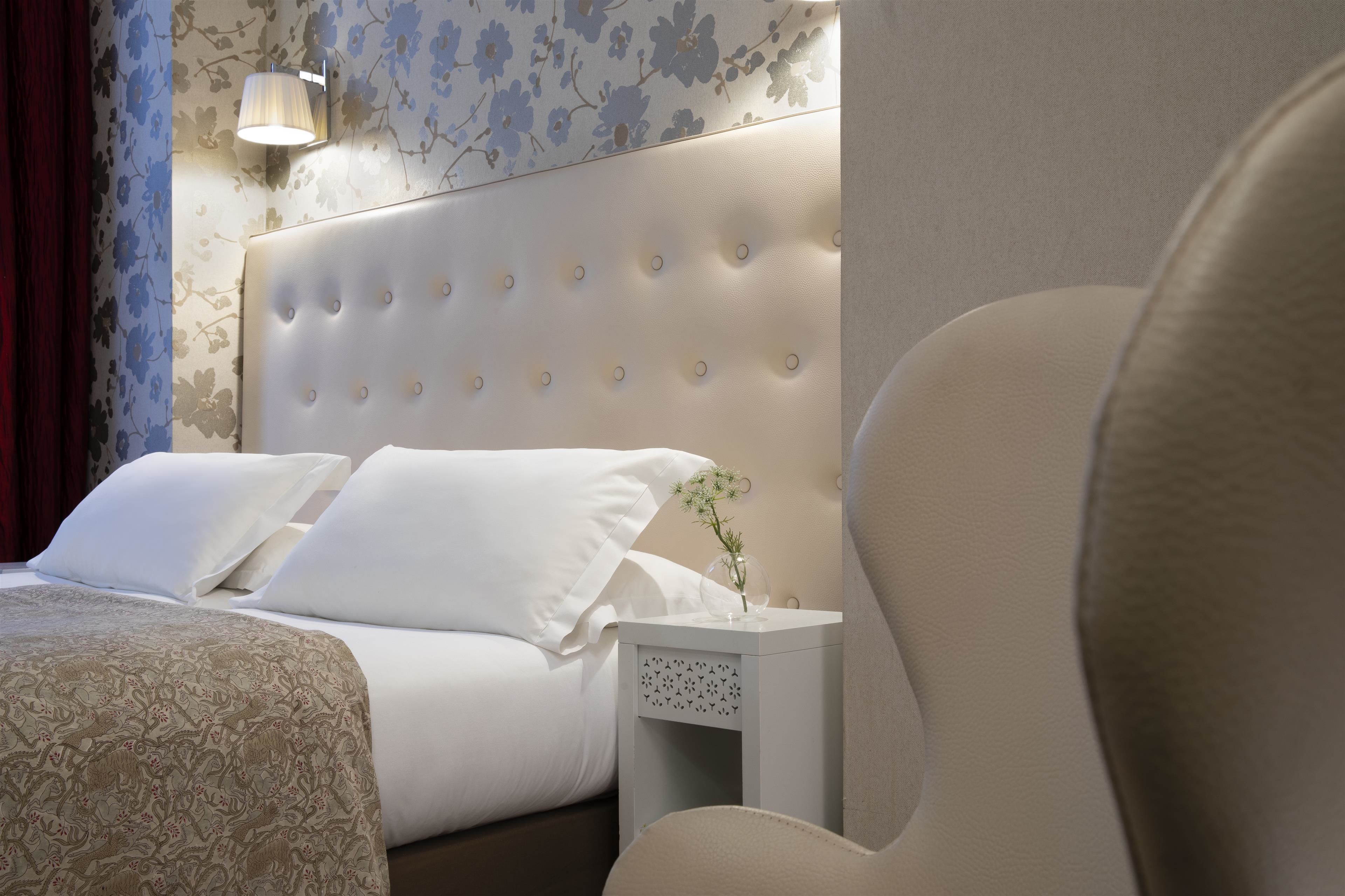 Un letto rifatto con lenzuola bianche e una coperta a motivi floreali, una testiera imbottita, un comodino con un vaso di fiori e una parete con carta da parati floreale.