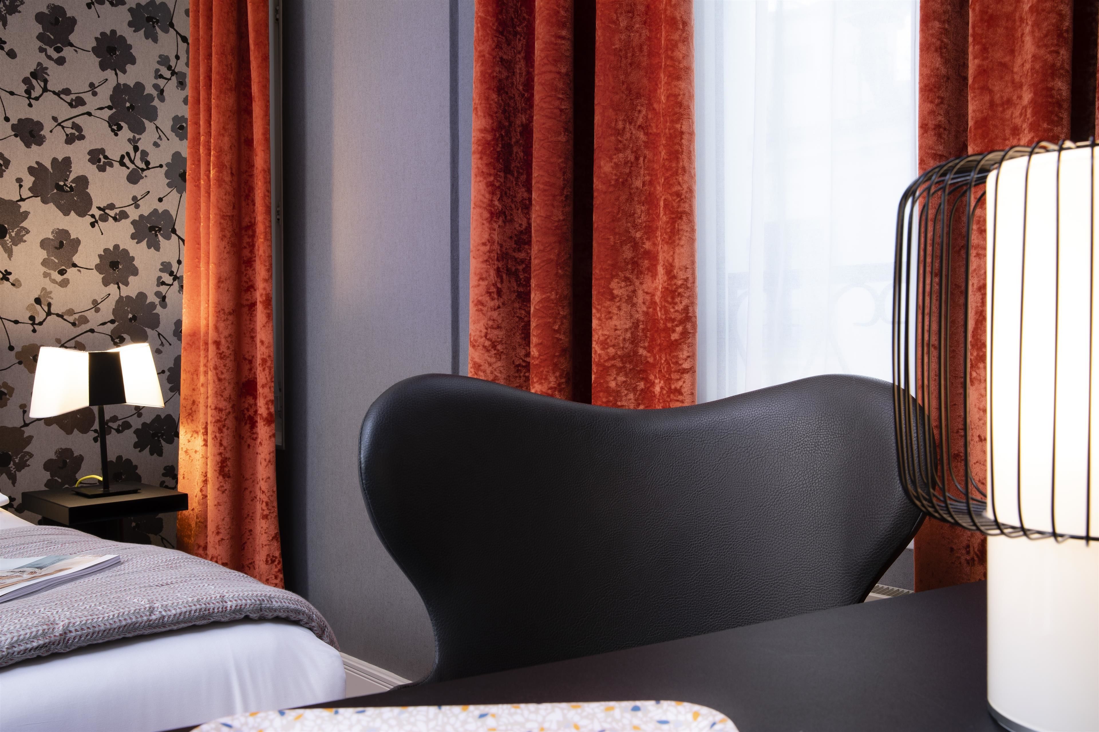 Une chambre d’hôtel avec un lit, un bureau, une chaise, une lampe et des rideaux rouges.