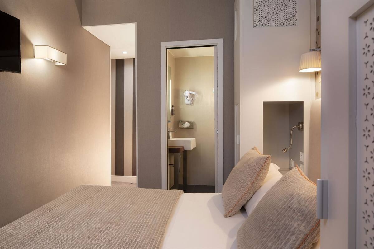 Une chambre à coucher avec un lit, une table de chevet, un téléviseur fixé au mur et une salle de bain visible à travers une porte ouverte.