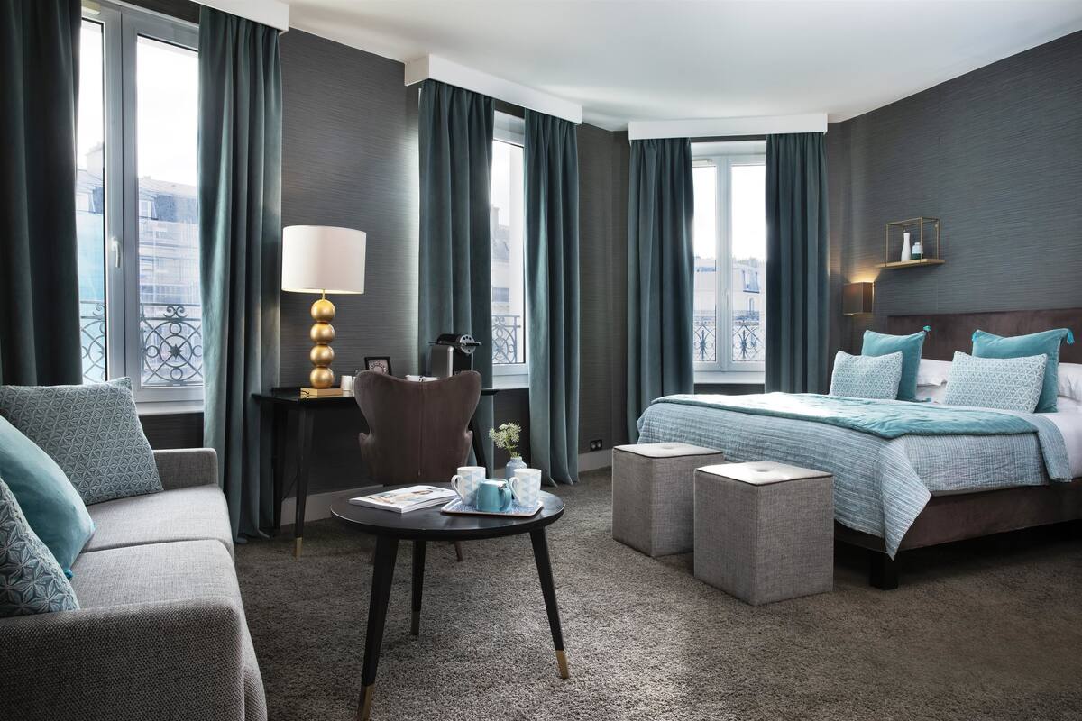 Une chambre d’hôtel moderne, équipée d’un lit, d’un canapé, d’une petite table avec une lampe, et offrant une vue sur la ville grâce à de grandes fenêtres.