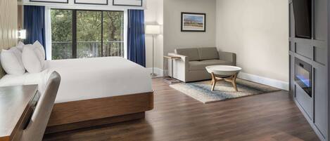 Habitación de hotel con una cama grande, un sofá, una mesita, un televisor y un balcón con vistas a árboles.