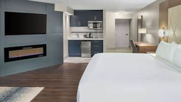 A modern hotel room with a large bed, a built-in TV, a kitchen area, and a seating space.
