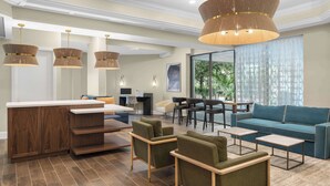 Un hall d’hôtel moderne avec un comptoir d’accueil en bois, des fauteuils verts, un canapé bleu et une grande fenêtre donnant sur un paysage verdoyant.