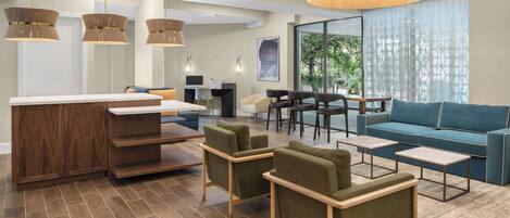 A modern hotel lobby with a wooden reception desk, green armchairs, a blue sofa, and a large window offering a view of greenery.