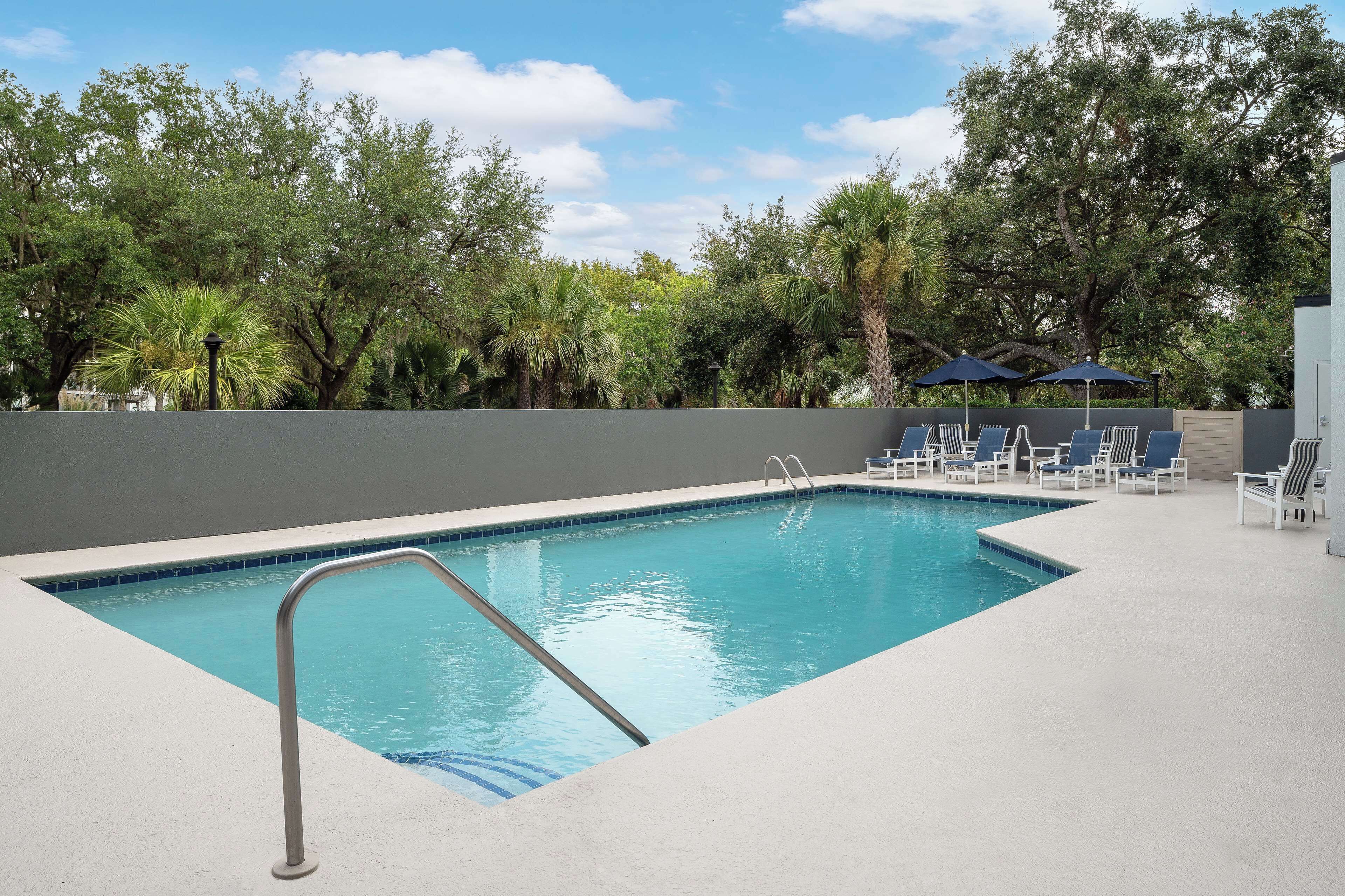 A rectangular swimming pool with a metal ladder, surrounded by a light-colored deck. There are lounge chairs and umbrellas in the background.