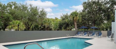 Una piscina rectangular con una escalera metálica, rodeada por un deck de color claro. Se observan sillas de descanso y sombrillas al fondo.
