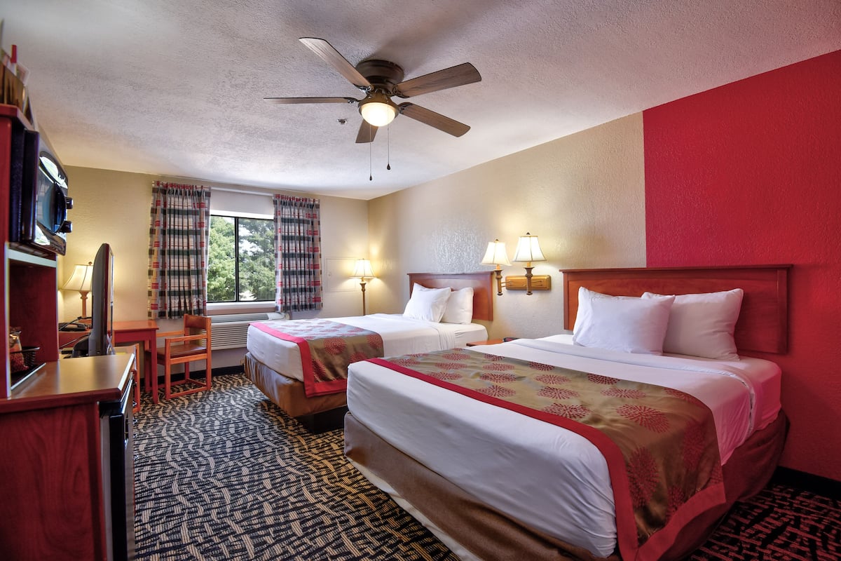 A hotel room with two beds, a desk, a chair, a ceiling fan, and a window with curtains.