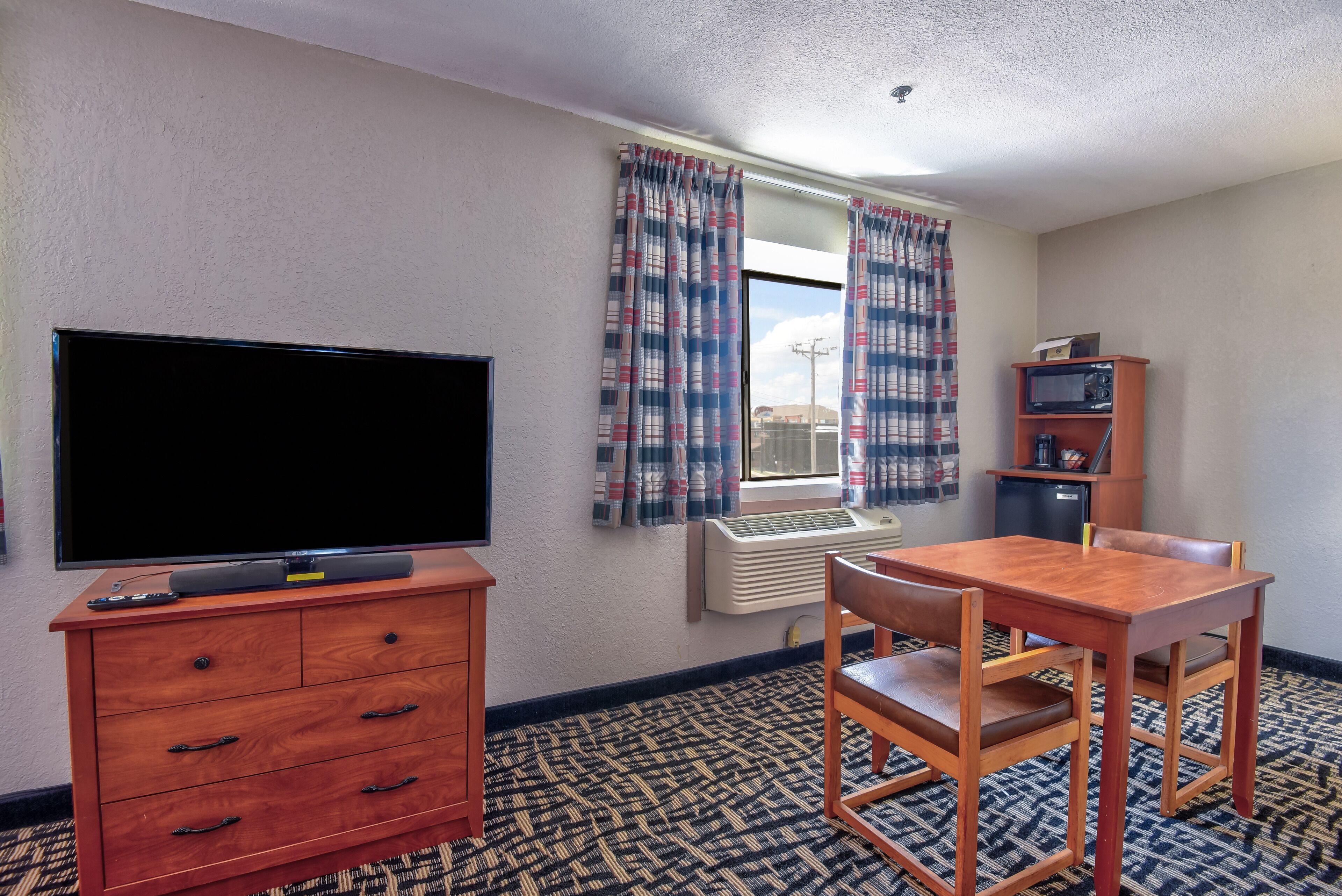 Habitación de hotel con televisor de pantalla plana, cómoda de madera, mesa de comedor con sillas, microondas y ventana con cortinas a cuadros.