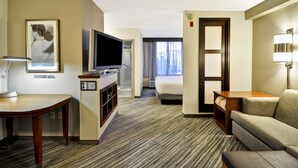 A hotel room with a flat-screen TV, a desk with a lamp, a sofa, a bed, and a door with a light switch.