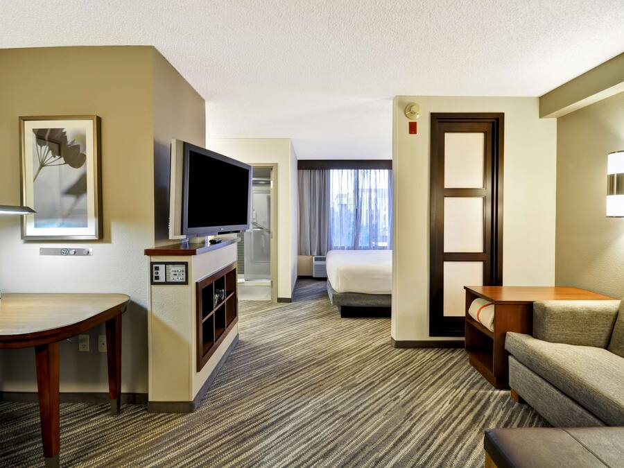 A hotel room with a flat-screen TV, a desk with a lamp, a sofa, a bed, and a door with a light switch.