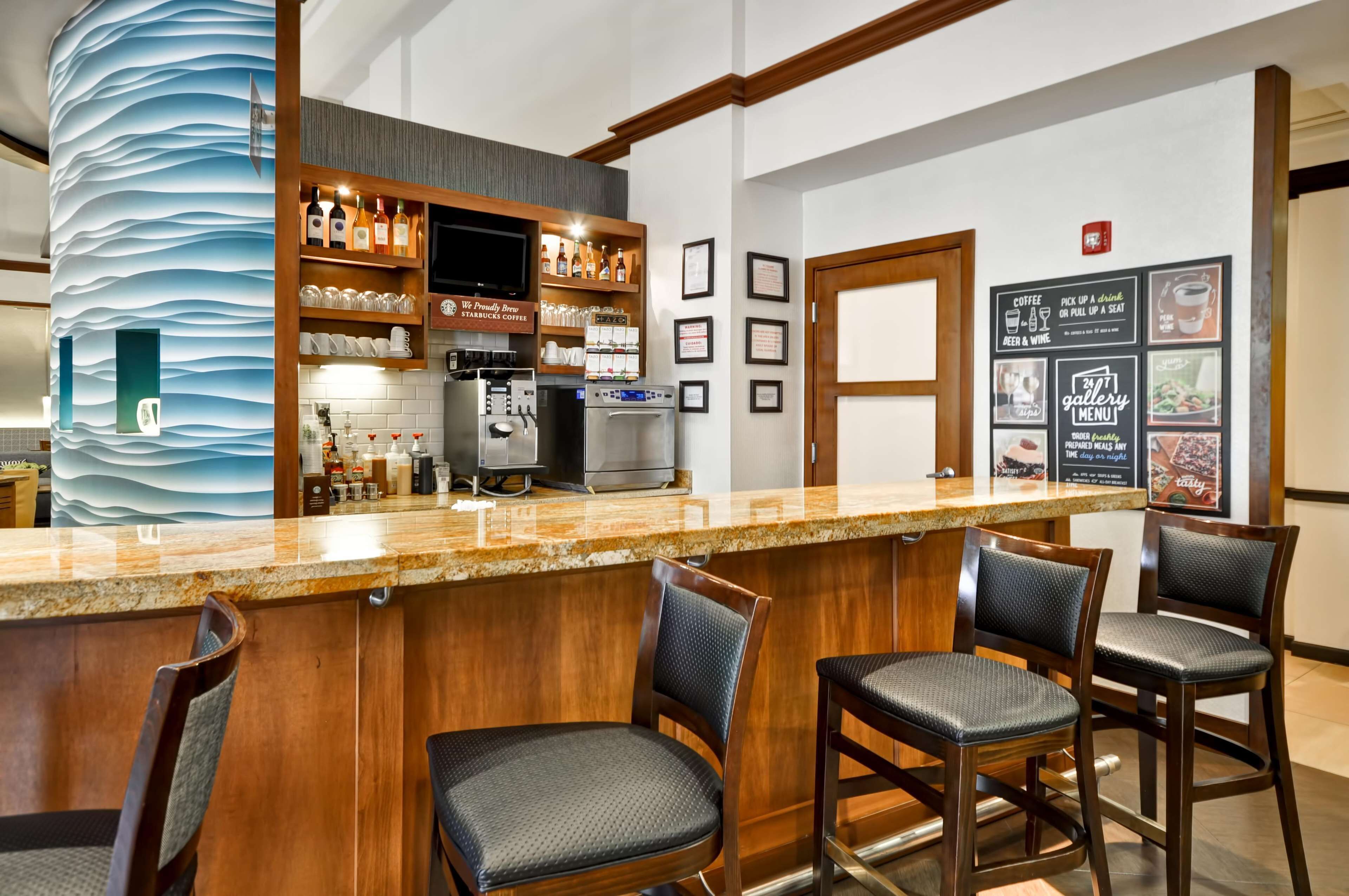 Una zona de bar con mostrador, taburetes, una máquina de café y estantes con botellas y vasos.