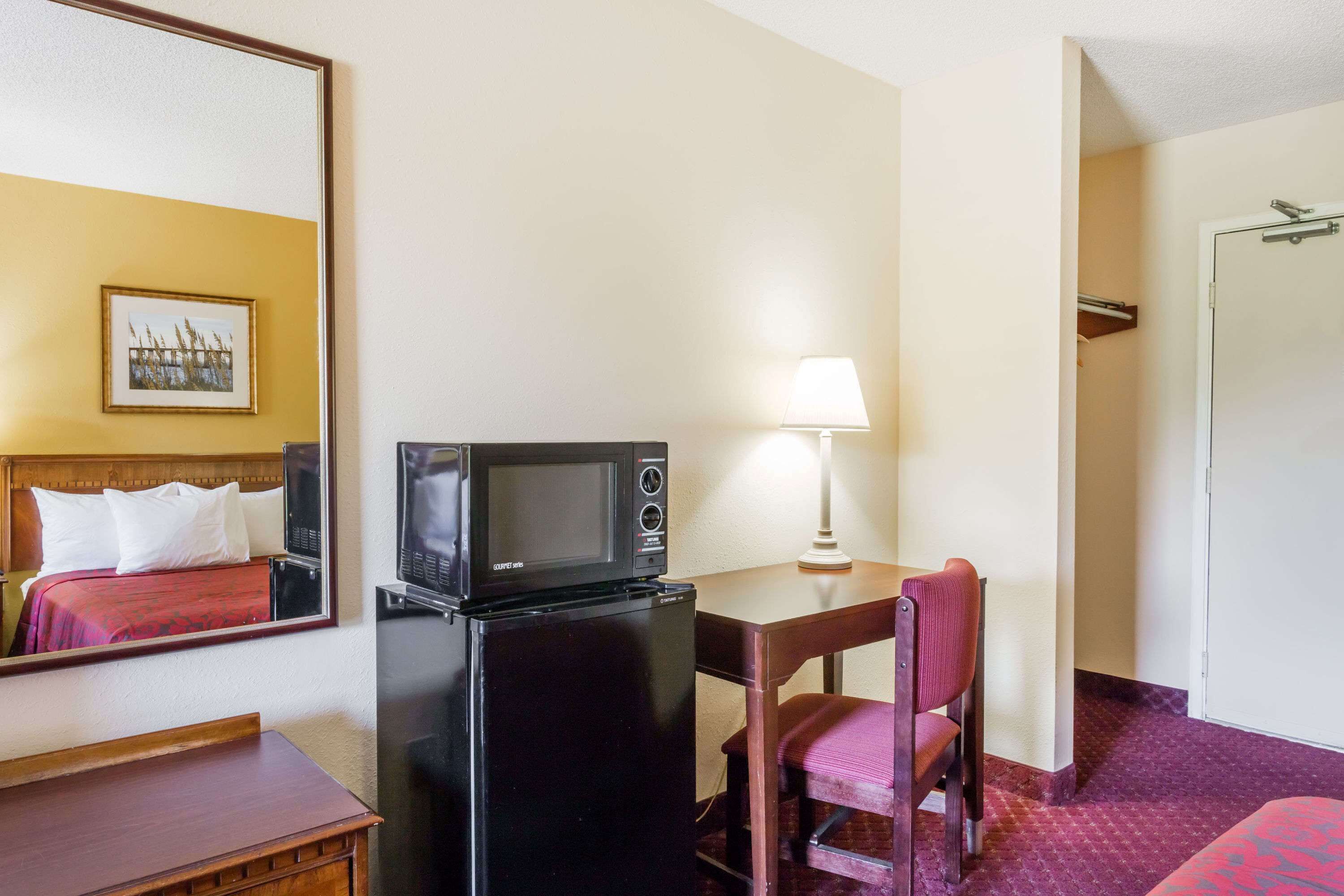 Een kleine hotelkamer met een bed, een klein bureau met een magnetron, een stoel en een lamp.