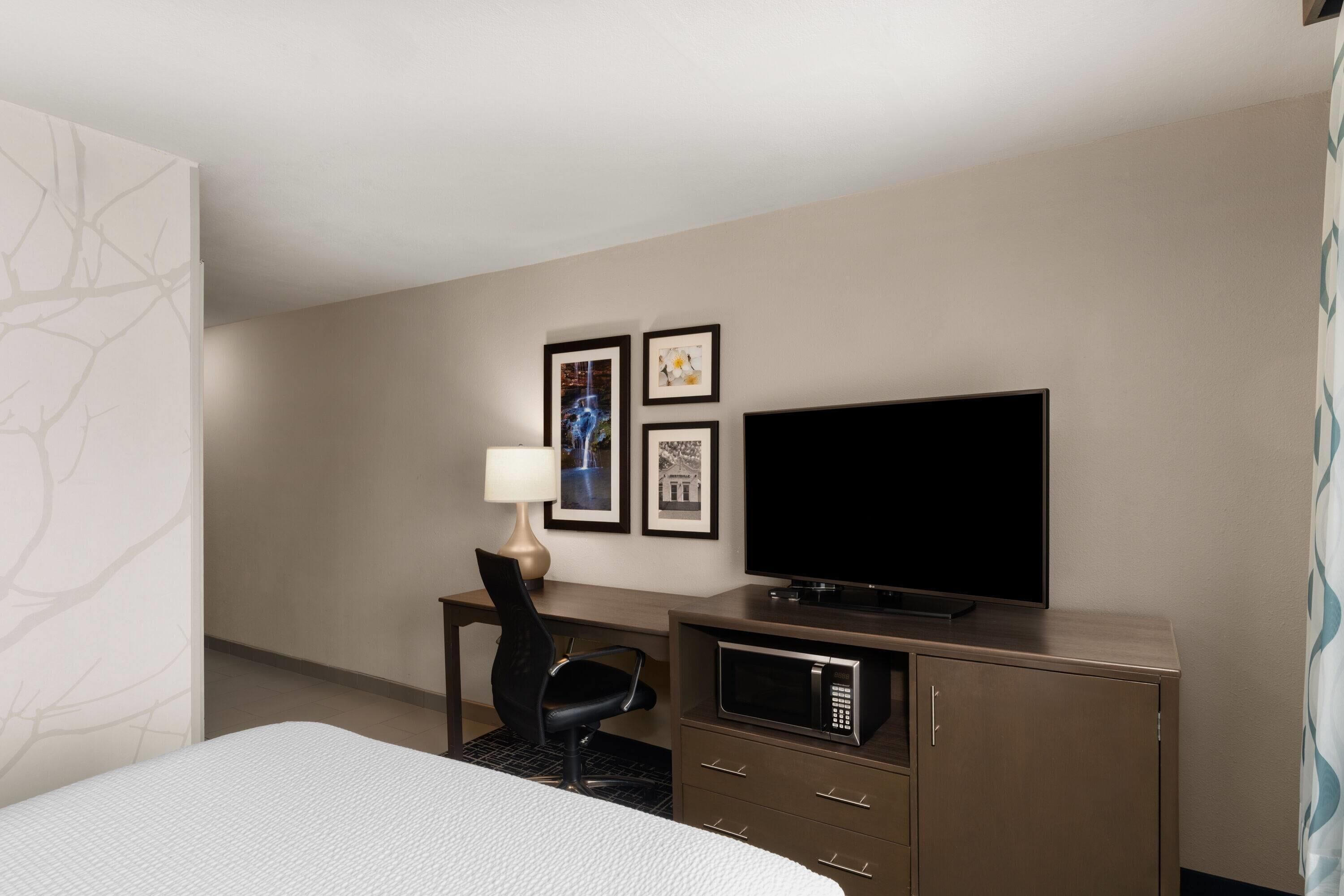 A hotel room with a bed, a desk with a chair, a flat-screen TV, a microwave, and framed pictures on the wall.