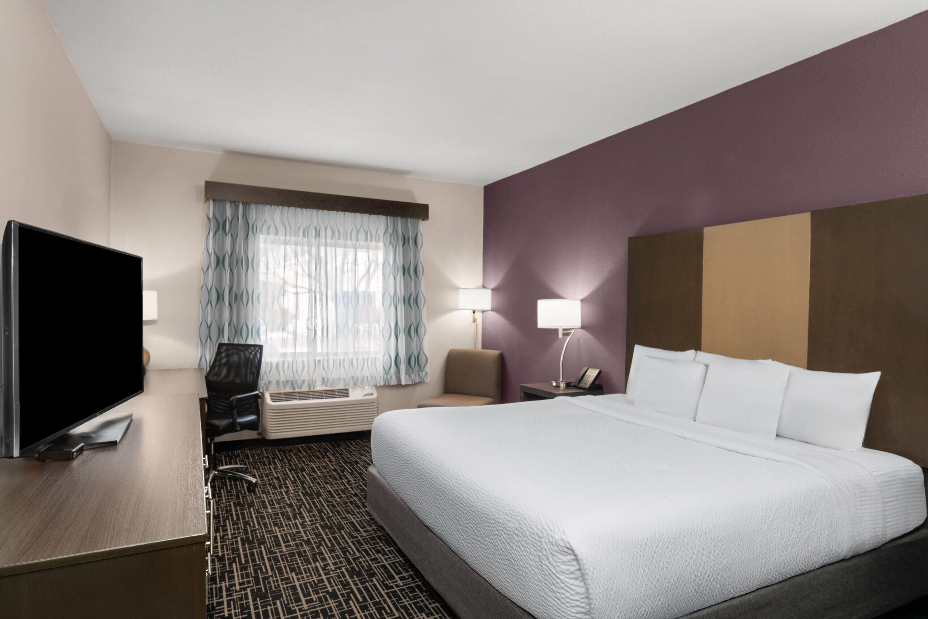 A hotel room with a bed, a desk with a TV, a chair, a lamp, and a window with curtains.