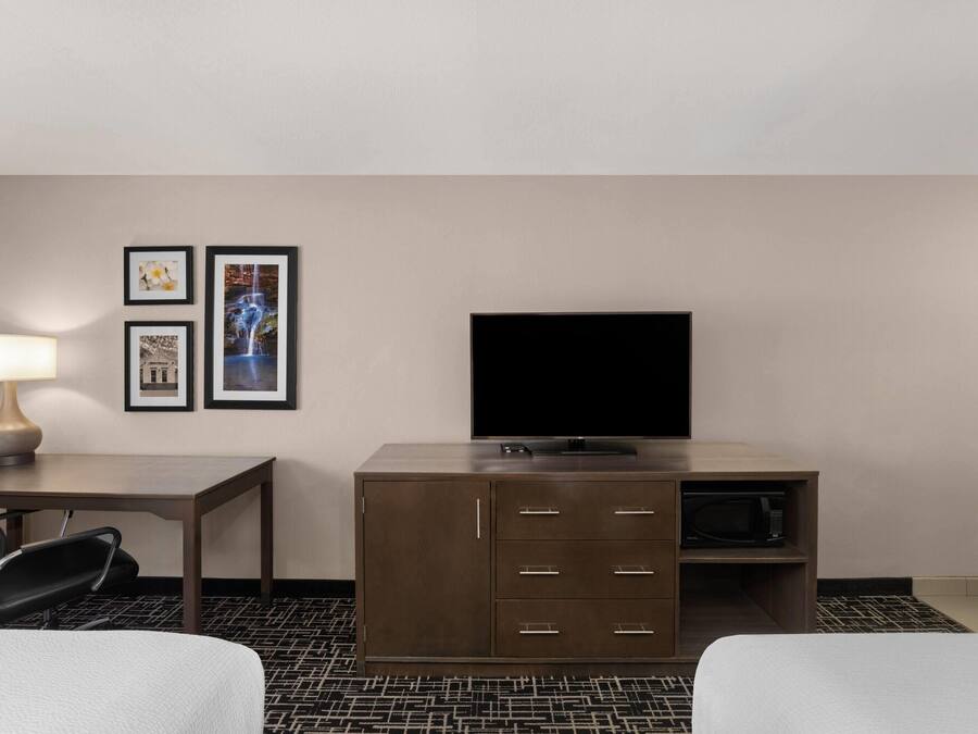Hotellihuone, jossa on sänky, kirjoituspöytä lampun kanssa, televisio, kaappi ja kaksi kehystettyä kuvaa seinällä.