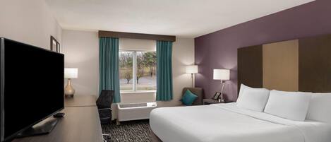 A hotel room with a bed, a TV, a desk, a chair, a window with curtains, and a lamp.