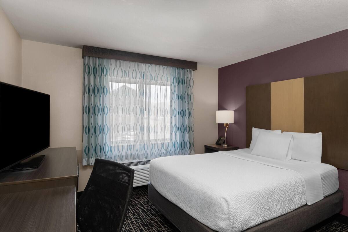 Une chambre d’hôtel avec un lit, un bureau avec une télévision, une chaise, une lampe et une fenêtre avec des rideaux.