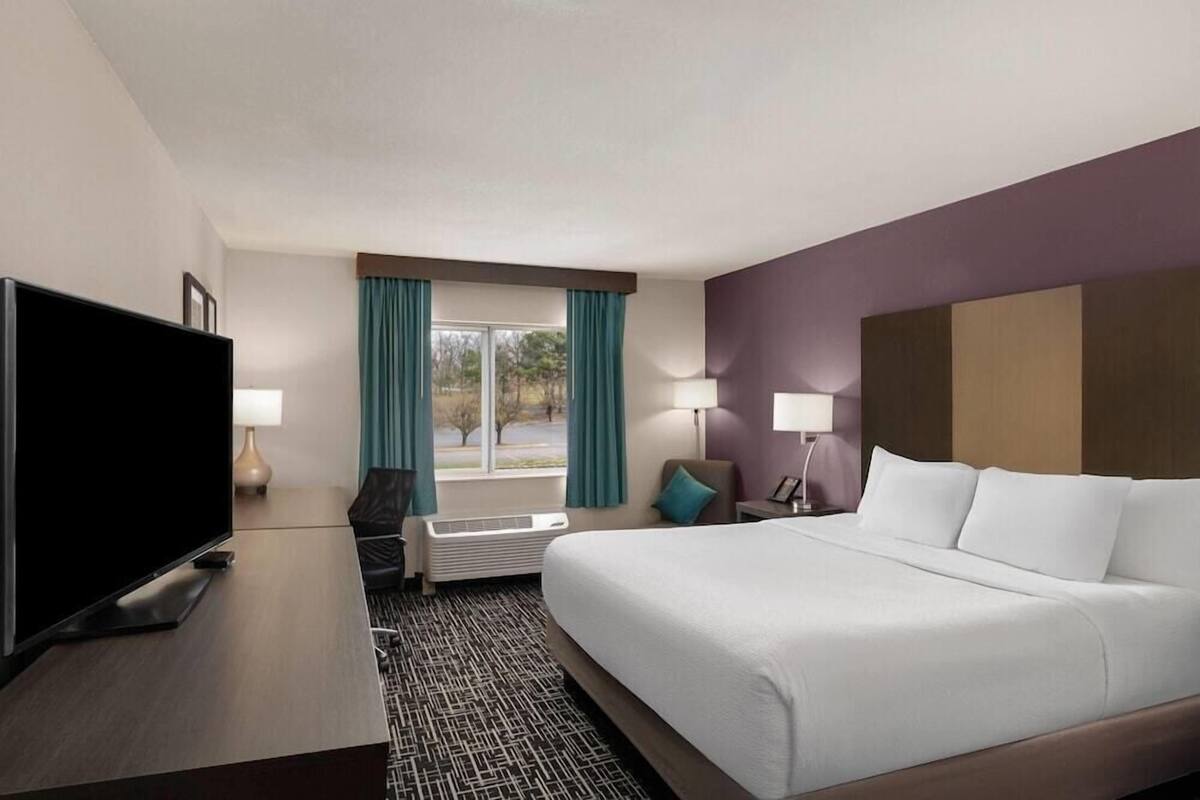 Une chambre d’hôtel comprenant un lit, une télévision, un bureau, une chaise, une fenêtre avec des rideaux et une table de chevet avec une lampe.
