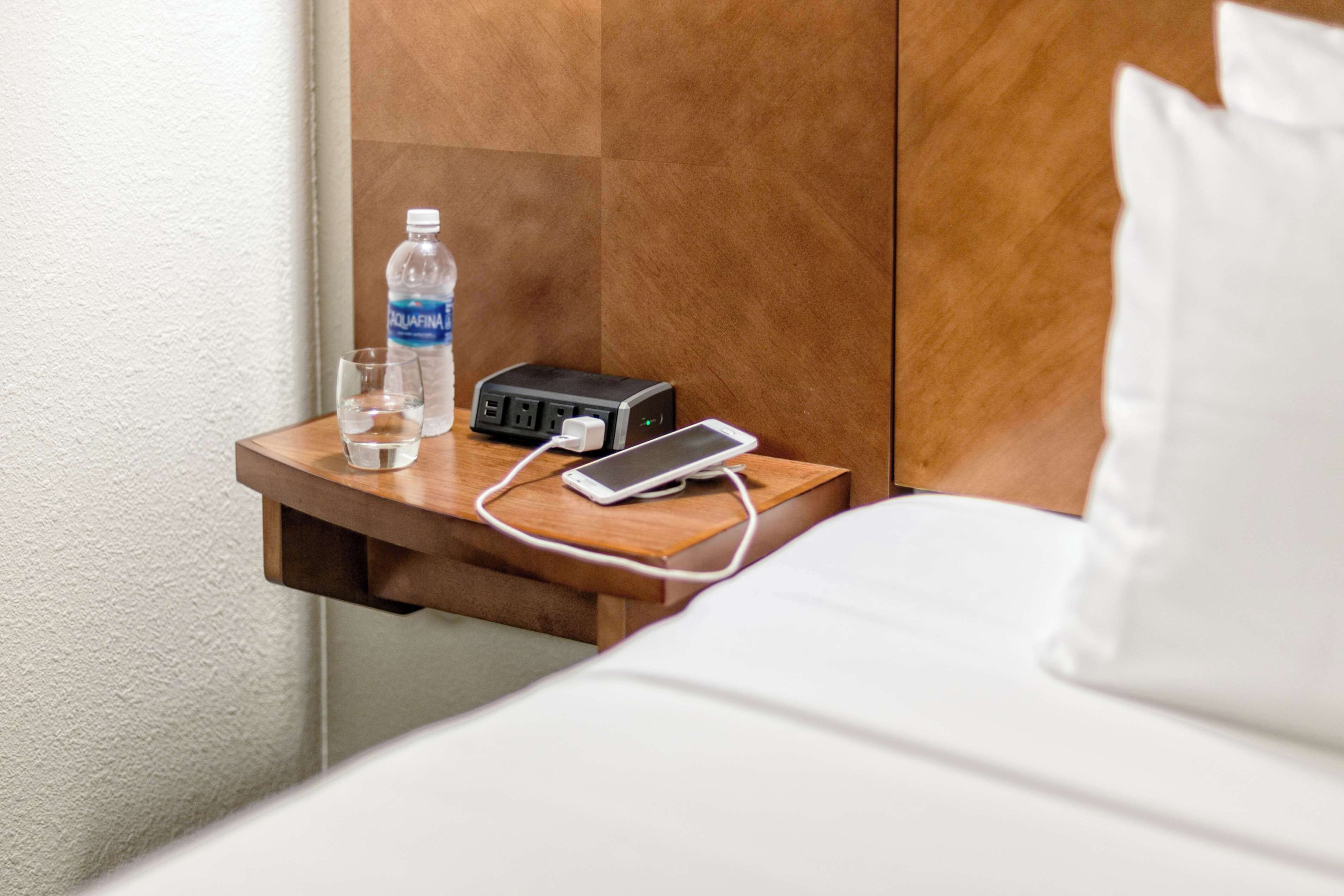 A hotel room with a wooden bedside table holding a water bottle, a glass, a power strip, and a smartphone charger.