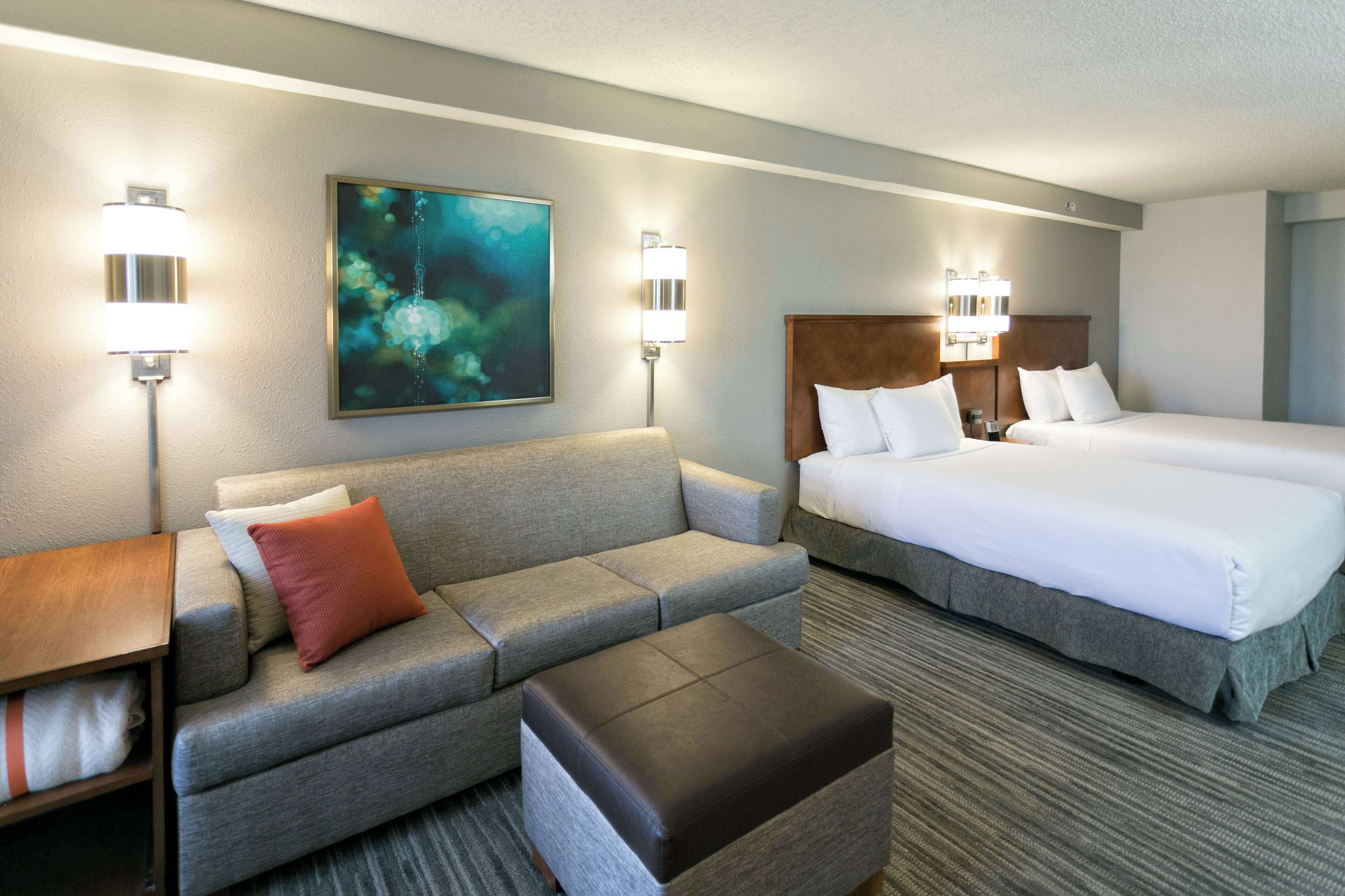 Quarto de hotel com sofá cinzento, um banquinho marrom, uma cama grande com roupa de cama branca e duas luminárias fixadas na parede.