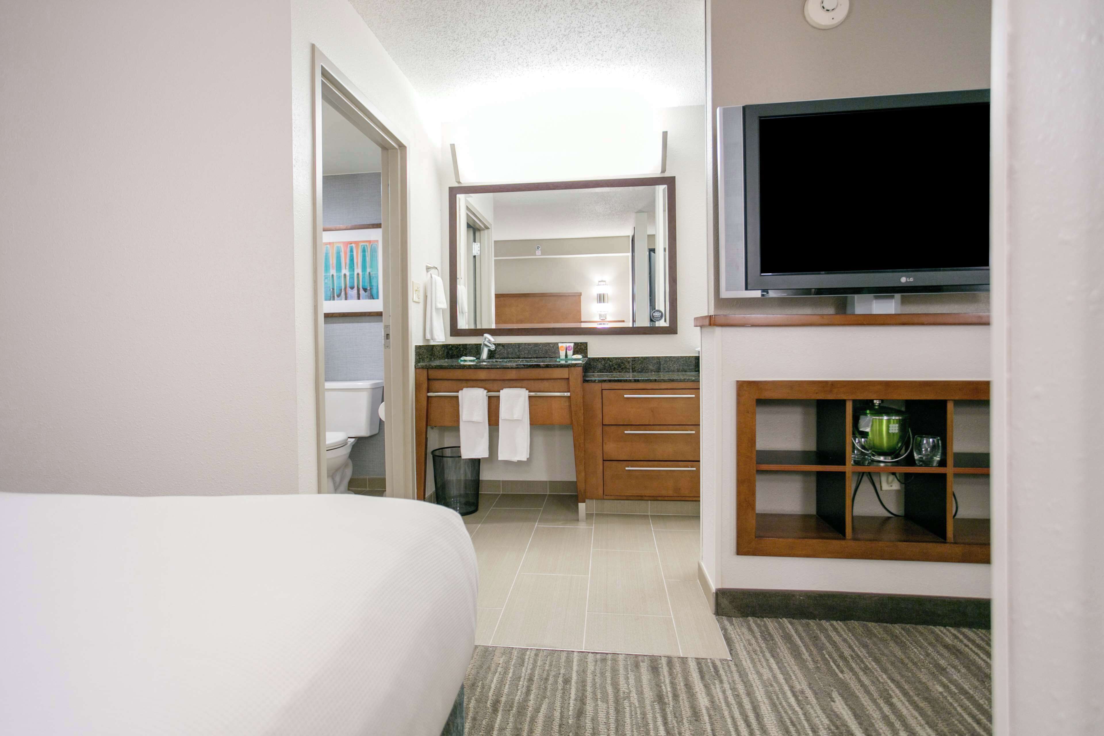 Habitación de hotel con cama, baño con lavamanos y espejo, y televisión en la pared.