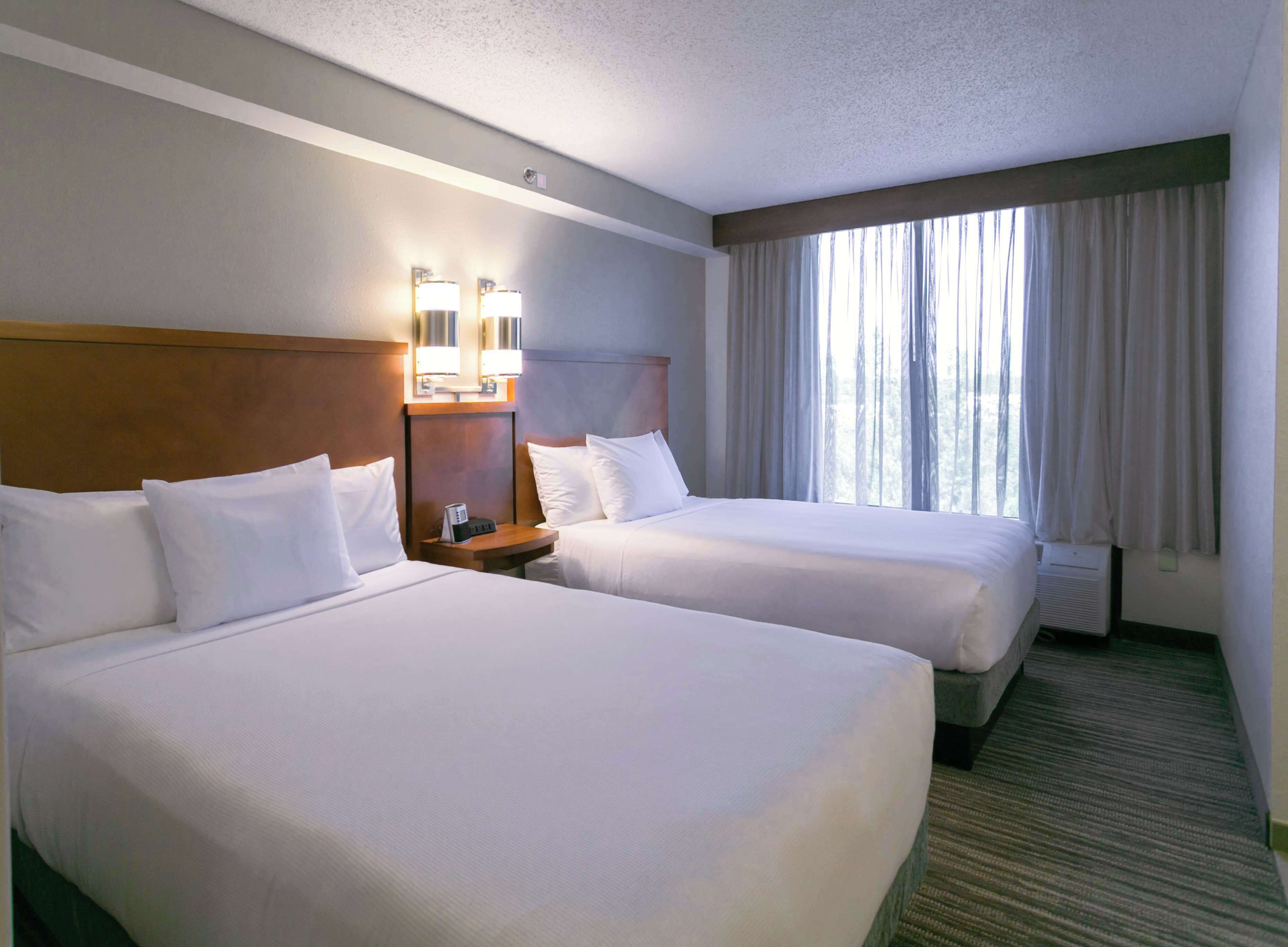 A hotel room with two beds, a nightstand with a lamp, and a large window with curtains.