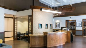 A modern lobby with a central wooden reception desk, seating area, and a kitchen in the background. - Hyatt Place Nashville/Franklin/Cool Springs (Franklin)