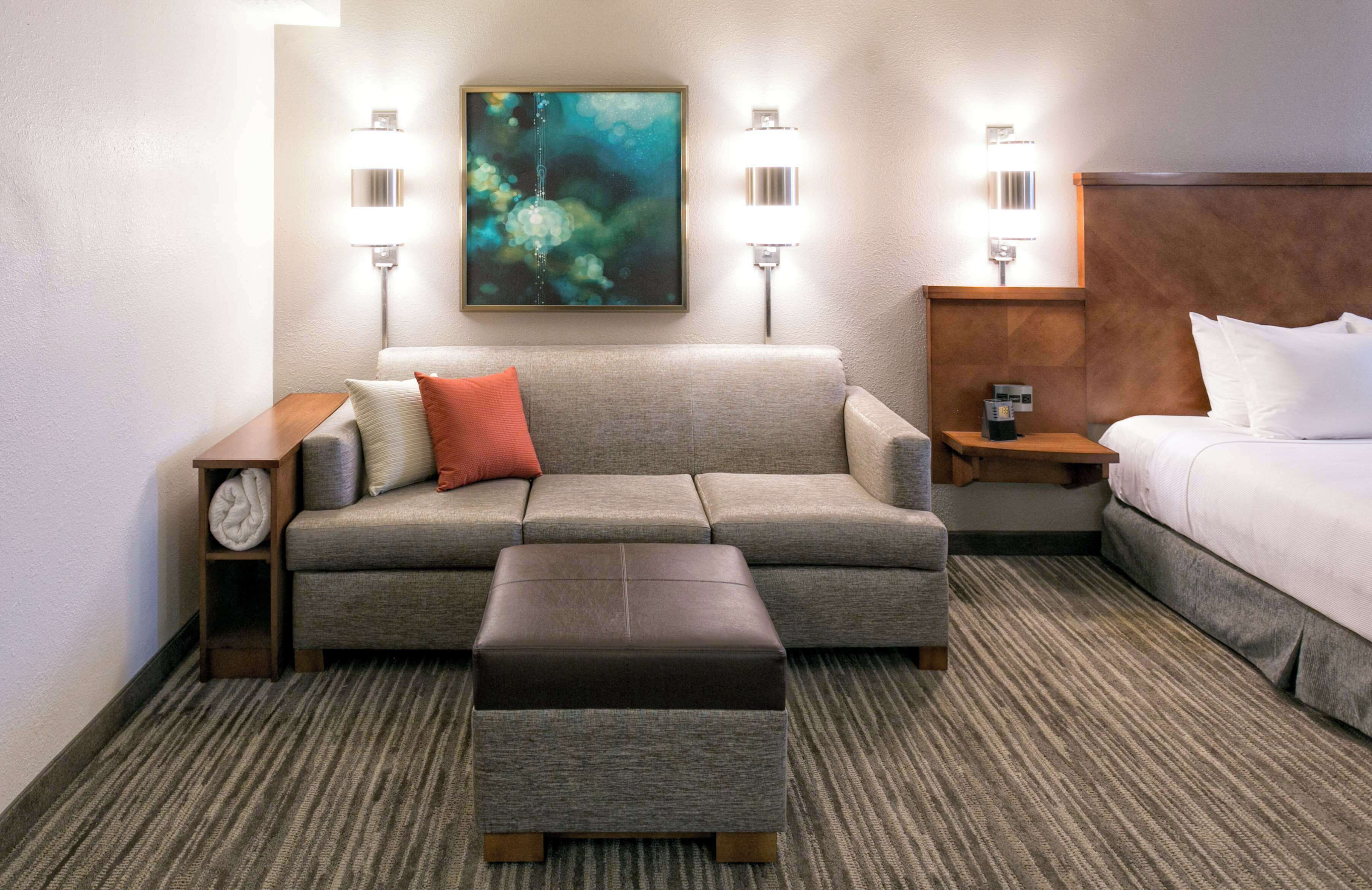 Ein Hotelzimmer mit grauem Sofa, einem kleinen Hocker, einem Bett mit weißen Leinen und einem Gemälde an der Wand.