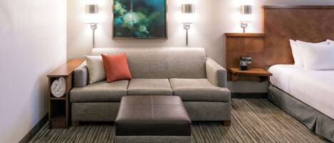 Ein Hotelzimmer mit grauem Sofa, einem kleinen Hocker, einem Bett mit weißen Leinen und einem Gemälde an der Wand.