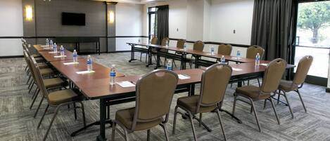 Un salón de conferencias con una larga mesa, sillas, botellas de agua y un televisor en la pared.